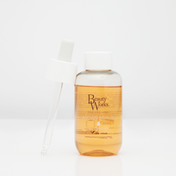 Beauty Works Argan Serum in a clear bottle showing golden-amber liquid, with a white dropper cap and glass pipette resting beside it on a white background.