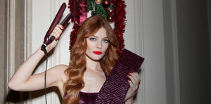 Woman with long red hair holding a hair straightener and a decorative ribbon against a neutral background.